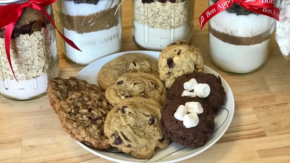 A plate of four kinds of cookies sits on a wood counter in front of jars of cookie mix tied with red ribbons.