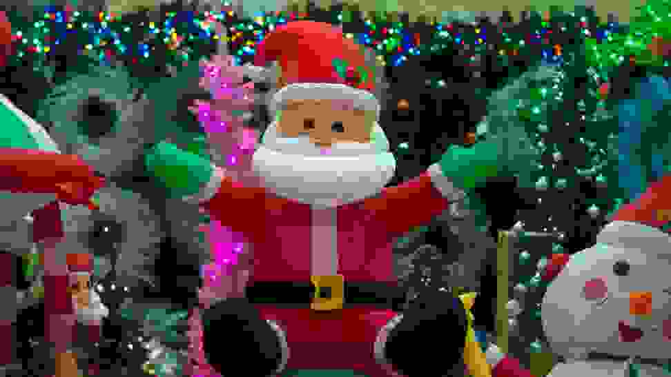 A light-up inflatable Santa decoration surrounded by other Christmas iconography: trees, colorful Christmas lights, an inflatable snowman, white wreaths, and more.