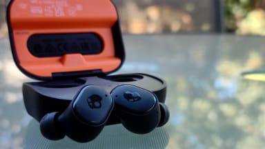 The SkullCandy Grind Fuel earbuds sit in all black with a skull logo along a glass-top table outside, their case open behind showing a bright orange interior.