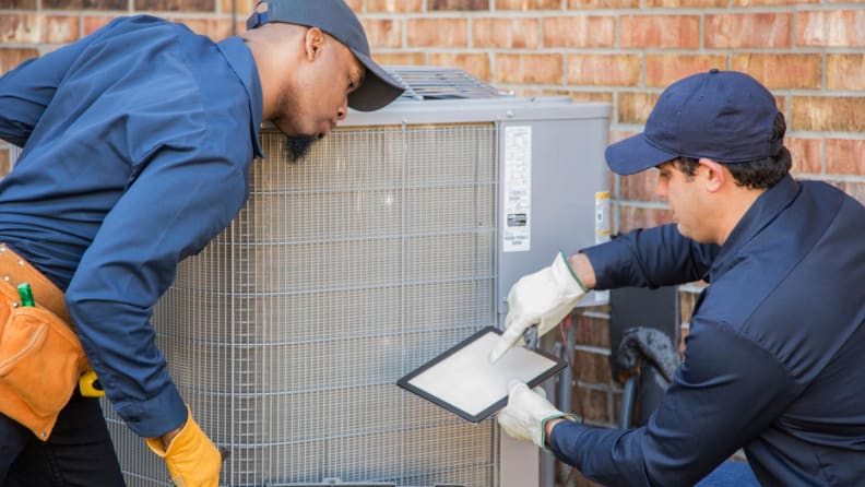 Two HVAC technicians stand in front of an outdoor air conditioner and look at a report.