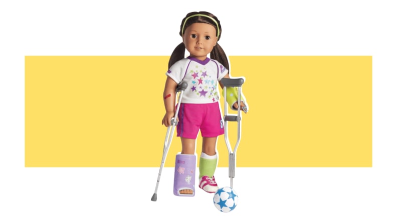 An American Girl doll with wearing athletic attire, with a purple foot cast on, and holding onto crutches.
