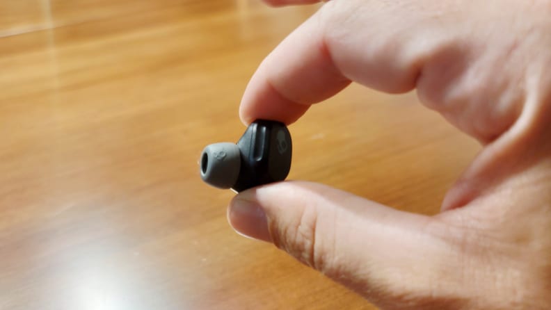 The all-black Skullcandy Mod earbud sits between thumb and finger above a wooden cabinet.