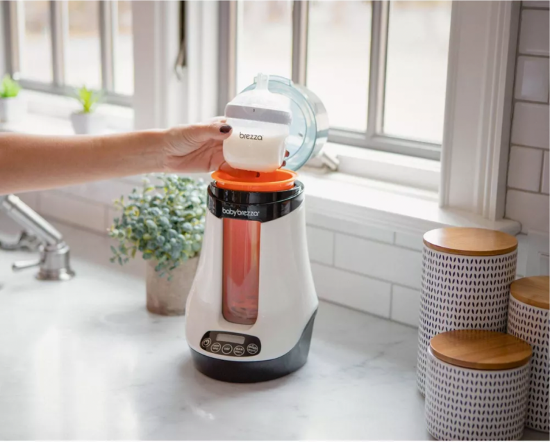 A hand holds a full bottle over the opening to the Baby Brezza Electric Bottle Warmer, which is sitting on a white countertop.