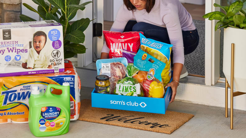 A woman brings a Sam’s Club grocery box filled with fresh produce and pantry staples inside her home, surrounded by bulk household items on the doorstep.