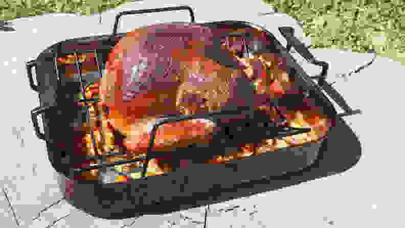 A smoked turkey sits in a roasting pan, the base of which is filled with veggies and other aromatics. The roasting pan is placed on pavement in the afternoon sun.