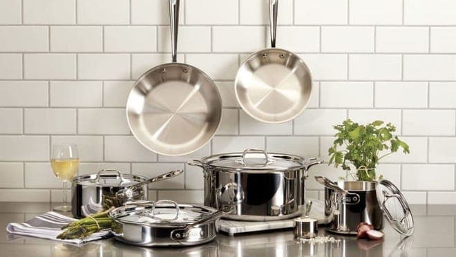 Silver pans hanging against a brickwall backsplash with assorted metal pots on a counter spread.