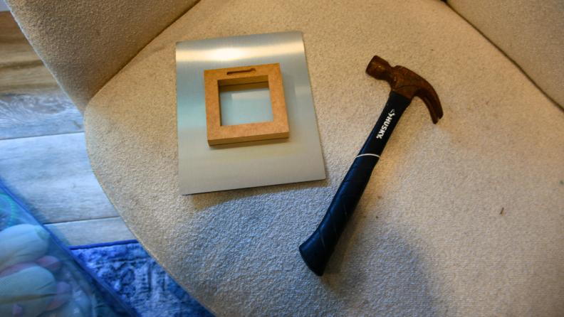 A hammer and metal photo with the hanging bracket showing from Frameology are sitting on a white chair.