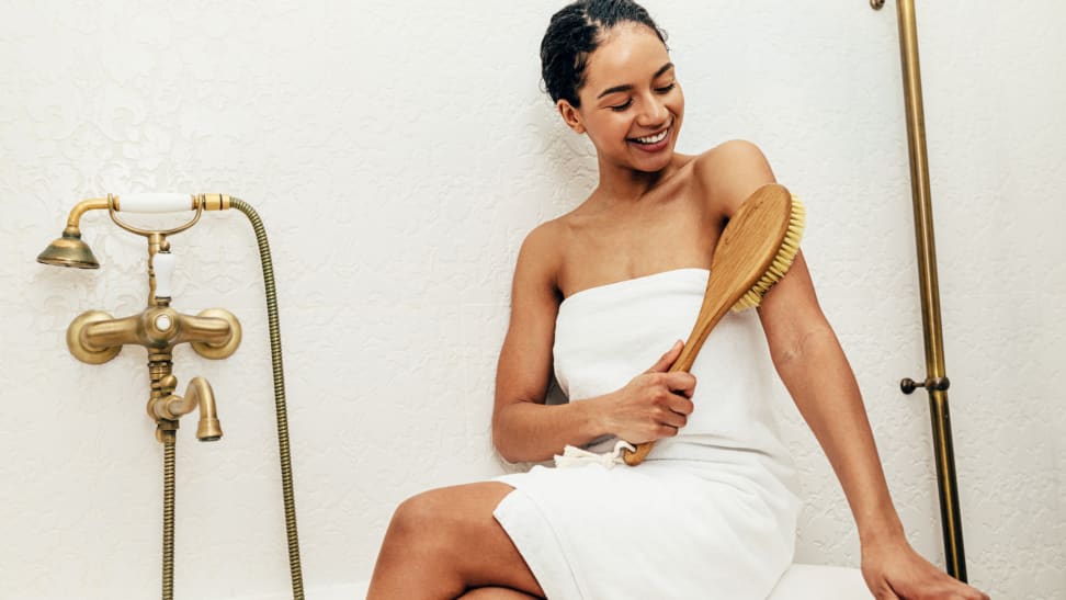 A woman sitting on the edge of a tub dry brushing her arm