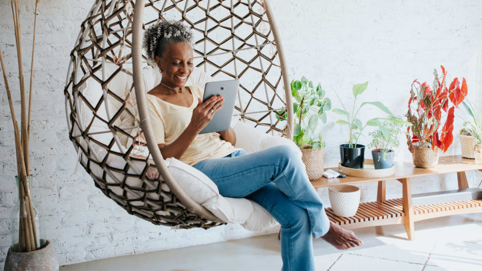 A person sits on a swing chair with a tablet in their home.