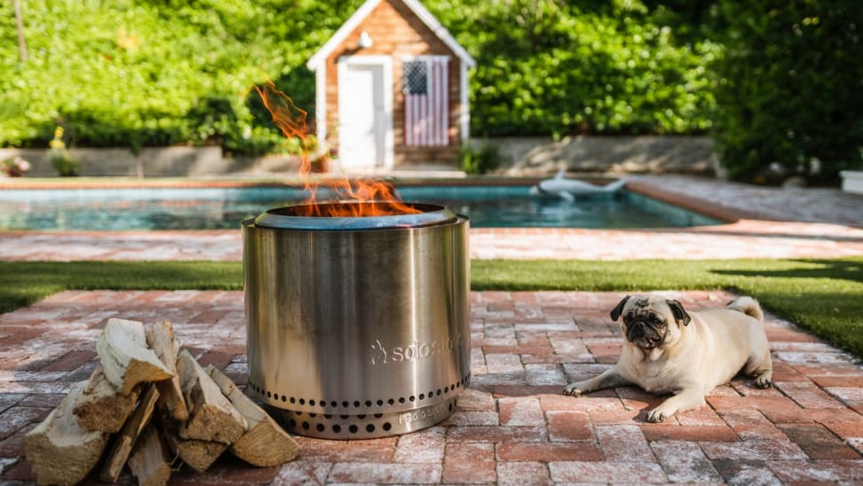 A dog sits to the right of a lit Solo Stove in a backyard.