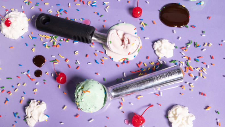 Las cucharas OXO y Zeroll llenas de helado yacen sobre un fondo morado rodeadas de chispas, cerezas y helado.