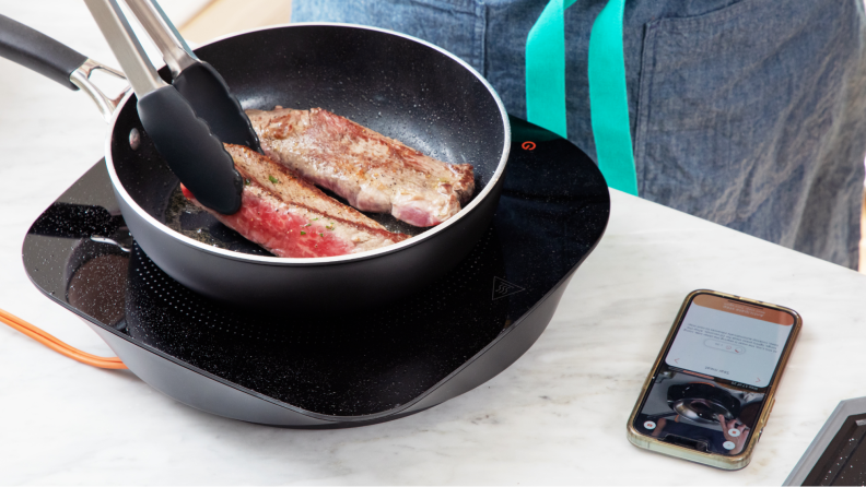 Two pieces of steak being cooked atop cookware with a smartphone displaying a recipe.