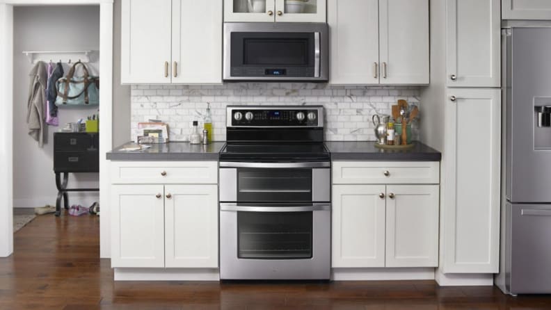 A lifestyle image of a modern home kitchen featuring a stainless steel double oven electric range framed by white cabinets.