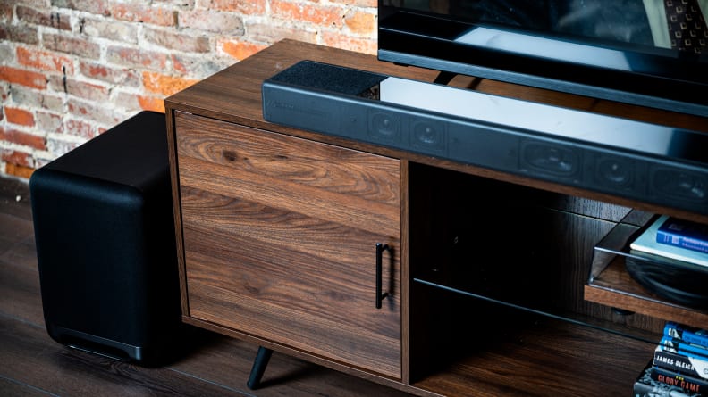 The all black Sony HT-A7000 sits atop a dark wood console against a brick wall below a large flat screen TV next to a large black subwoofer as shot from the right.