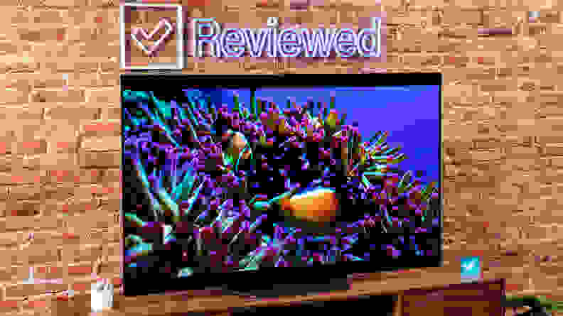 The LG B2 on a wooden table in front of a brick wall with a neon Reviewed sign displaying an ocean scene.