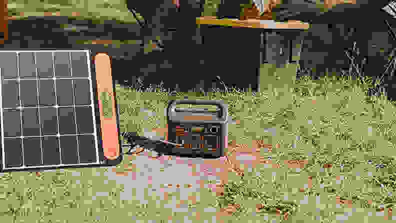 A power station sits on the ground in the woods, getting recharged by a nearby solar panel. In the background you can see two campers relaxing.