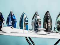 A line of steam irons sitting on top of an ironing board in front of a blue background.
