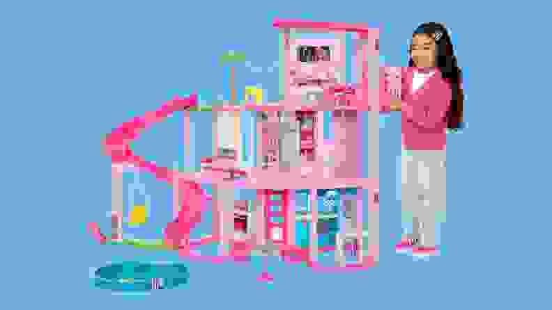 A young girl smiles as she stands next to the Barbie Dreamhouse 2023, with its three-story slide and pet play area.
