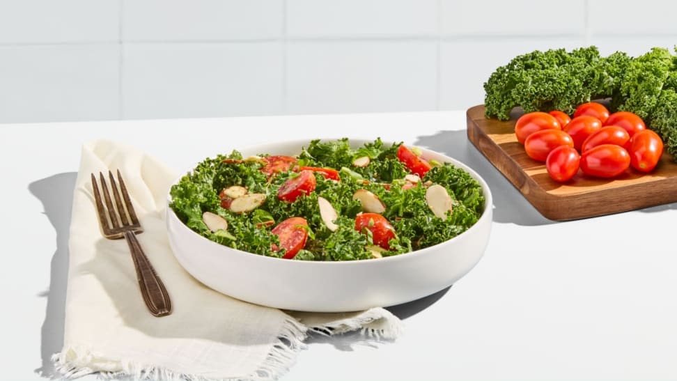 A bowl of salad on a white background next to a cutting board with kale and tomatoes.
