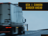 A truck drives a highway under a sign that reads USA Canada border