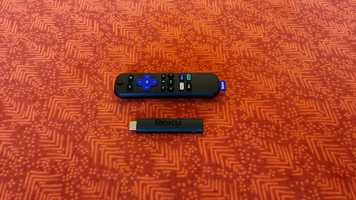 The matte black Roku Streaming Stick 4K is held in front of a brown TV console with a TV screen and speakers in the background.