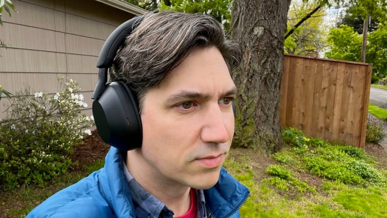 A man with brown and gray hair wears the matte black Sony WH-1000XM5 headphones in front of a large tree and a beige house.