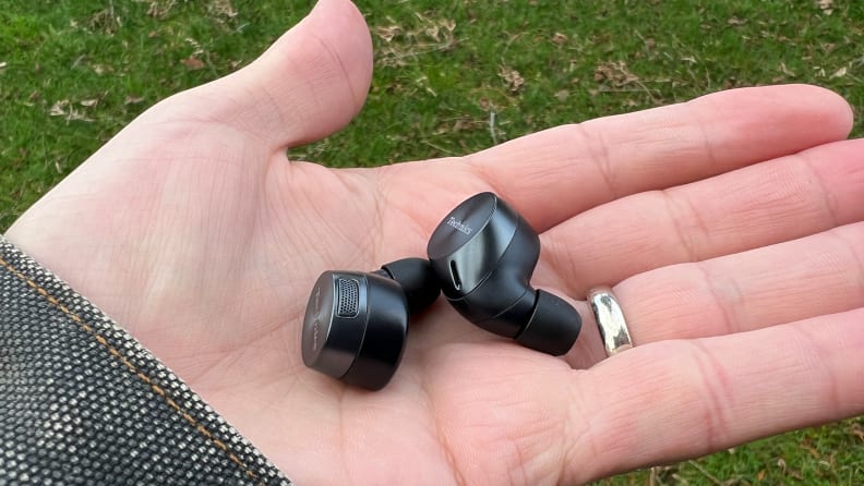 The gunsmoke colored wireless earbuds sit in a hand with a textured blue jacket and grass in the background.