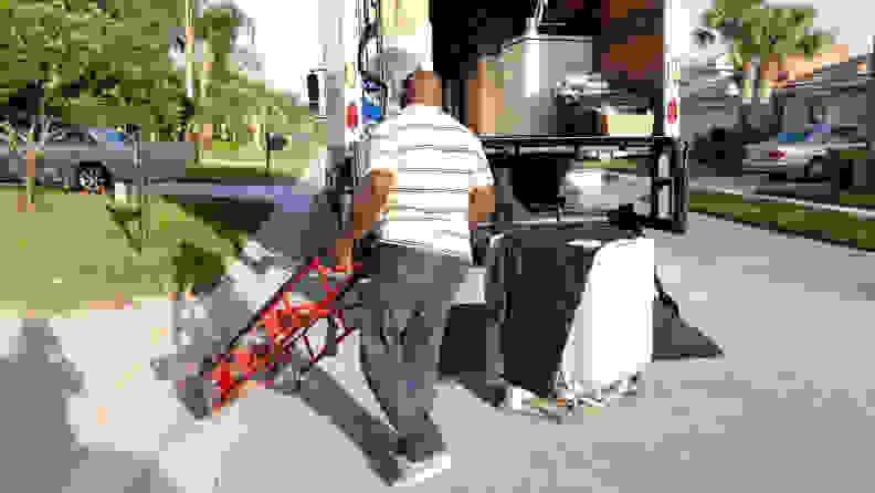 A man wheels a dolly over to an appliance that's sitting outside the open back of a delivery truck.