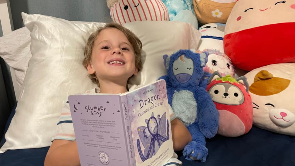 Small child smiling while laying down in bed against pillow next to Slumberkins Dragon Kin and other stuffed animals while holding book.