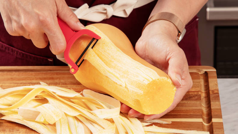 Person using Y-peeler tool to remove skin from butternut squash on top of wooden cutting board next to pile of skin.