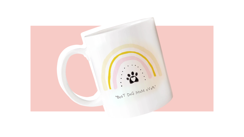 A white coffee mug with a warm colored rainbow above a black dog print, featuring text that says "Best dog mom ever", all against a pink background.