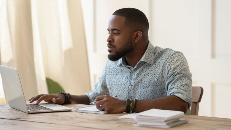 A man sits at a laptop, reading online appliance reviews and thoughtfully taking notes.