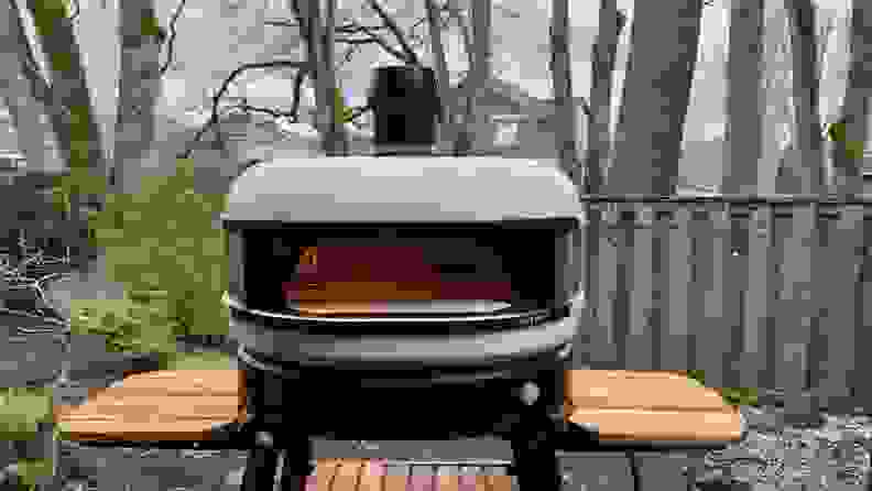 Gozney Dome Pizza Oven on stand with wood side tables, outdoors with open flame inside.