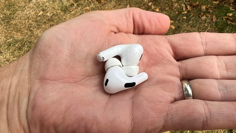 The white AirPods Pro earbuds sit in a hand with a wedding ring above leafy grass.