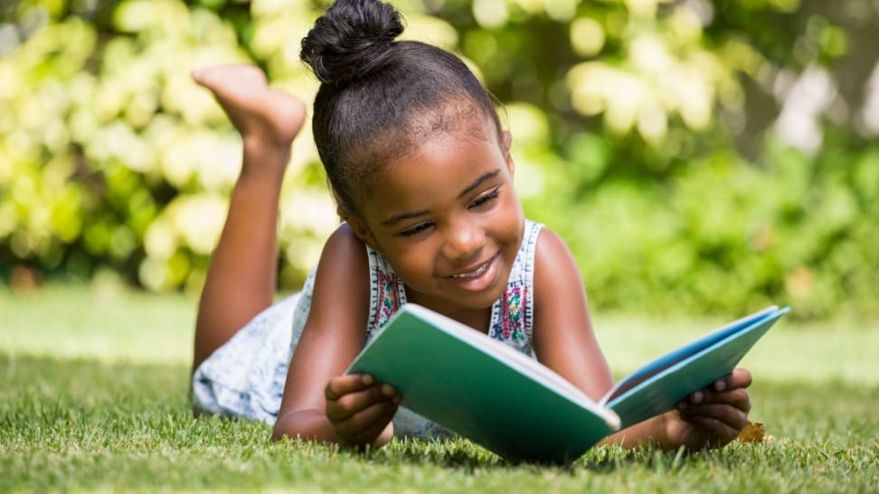 Young girl reading book outdoors in the grass while smiling