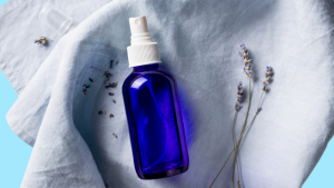 A blue bottle of pillow mist lies next to some lavender on a blue cloth, set on a blue background.