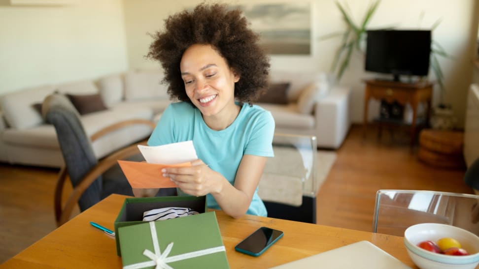 A person sits at a desk in a living room, smiling at a note from a care package in a green box.