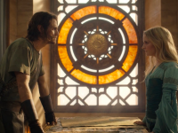 A still from 'The Rings of Power' that features Halbrand and Galadriel standing across fromo one another at a table, before a stained glass window.