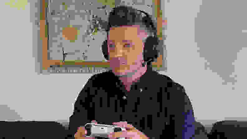 A Caucasian man with brown hair in a dark blue shirt sits on a couch in front of a painting while wearing the SteelSeries Arctis Nova Pro Wireless headset and holding a PlayStation 5 controller.