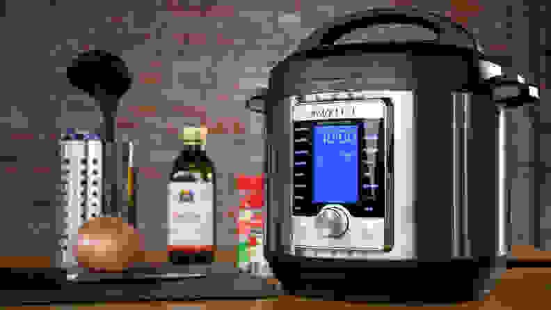 An Instant Pot sitting on a wooden counter top, with a whole onion on a cutting board beside it, along with seasoning bottles and olive oil.