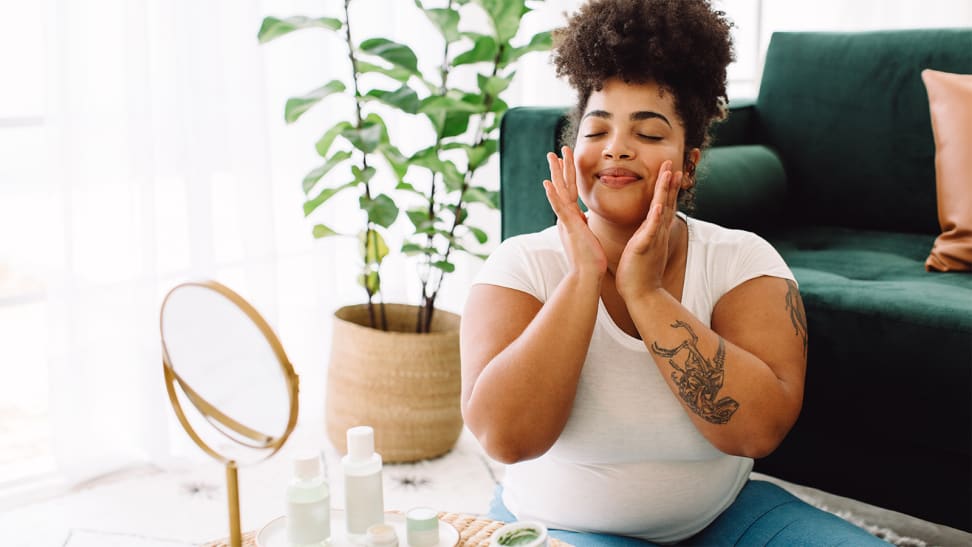 A person sits on the floor with a mirror and skincare items in front of them and touches their face.