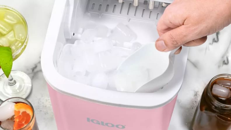 Igloo countertop ice maker with handle open as a person scoops ice from the basket beside mixed drinks.