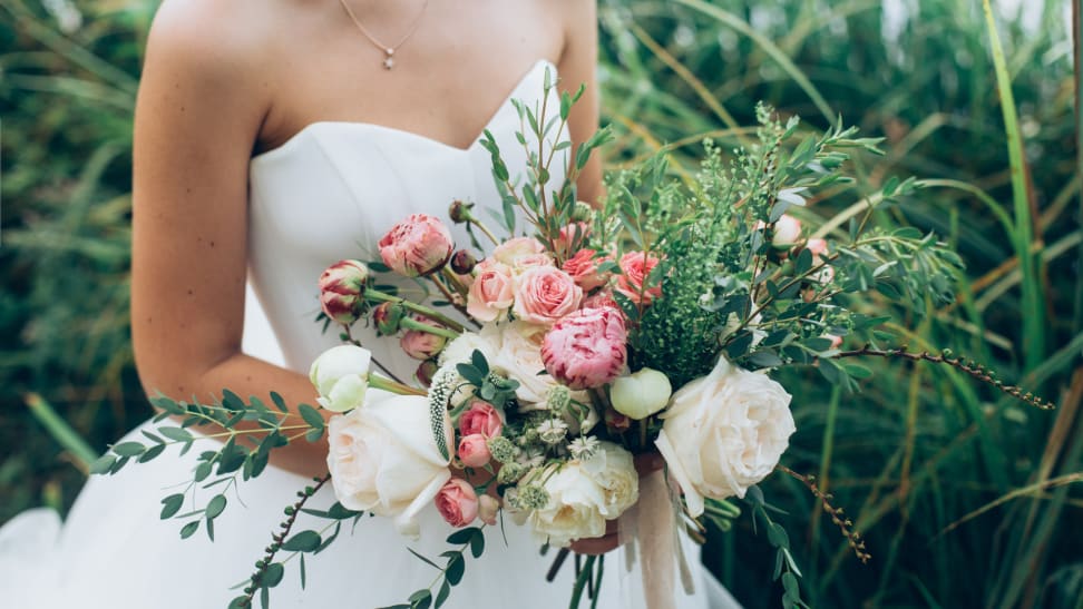 Bride in wedding dress holding colorful bouquet of rustic flowers outdoors.