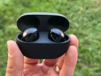 The Sony WF-1000XM5 in their charing case being held in someone's fingers above green grass.