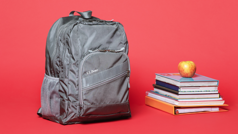 On a red background: A grey L.L. Bean Deluxe Book Pack backpack sitting next to a stack of books.