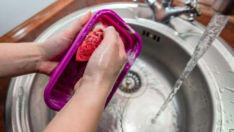 Hands scrub a pink sponge inside a plastic food container under running water in a kitchen sink.