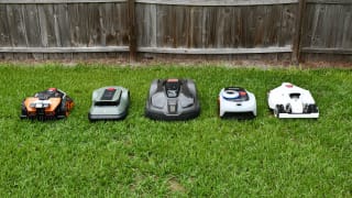 Five robot lawn mowers lined up together in the green grass with a fence behind