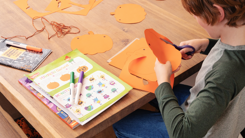 A child sits at a table and cuts out pumpkins from orange paper. Books and markets sit next to them.