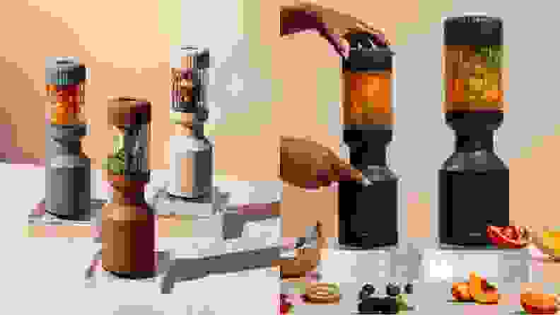 Photo collage of a blue, brown and cream Beast Blender Mini filled with different fruits next to a person's finger pressing button on a filled black Beast Blender Mini in order to activate blending while also next to a full size black Beast Blender Mini and surrounded by fruits.