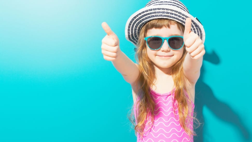 Little happy girl in beach outfit with hat and sunglasses holding thumbs up on blue background.